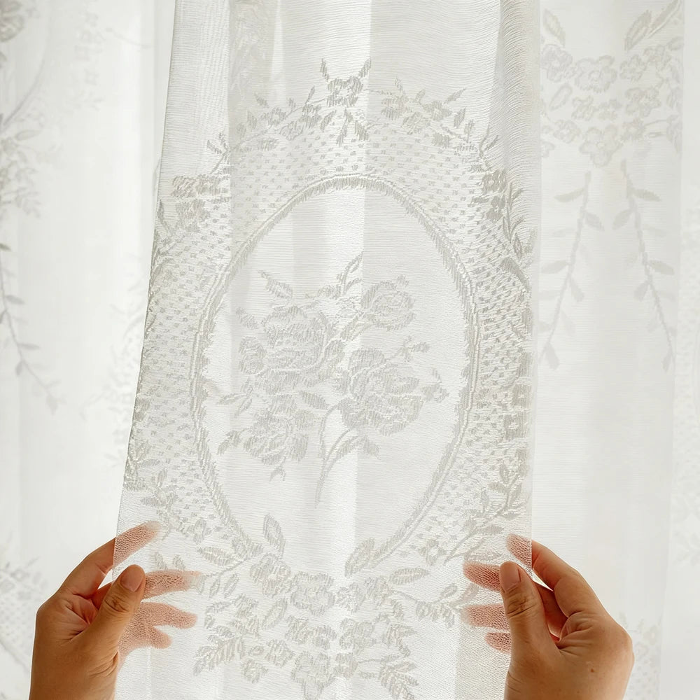 Hands holding a sheer white lace embroidered curtain with floral patterns.