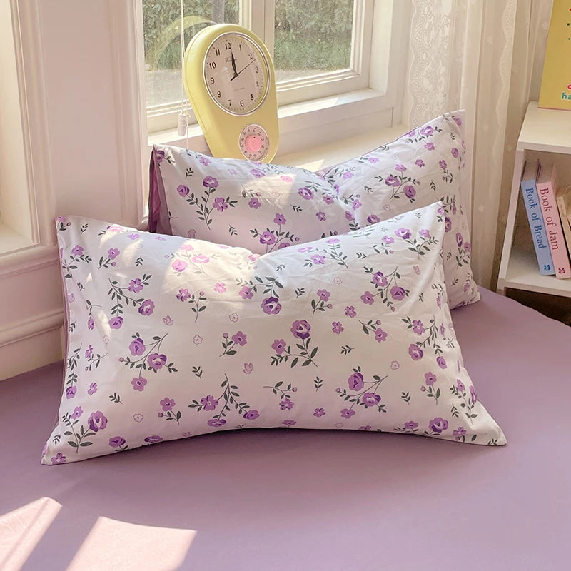 A set of white and purple floral patterned pillowcases and a yellow alarm clock displayed on a lilac surface.