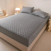 A gray waterproof quilted mattress protector is displayed on a bed, next to a lamp and a small white object.