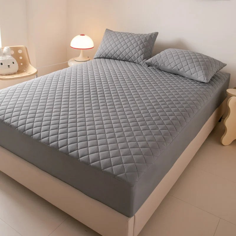 A gray waterproof quilted mattress protector is displayed on a bed, next to a lamp and a small white object.