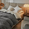 A cozy fleece blanket is draped over a light-colored bed with white pillows and a bedside lamp.