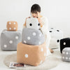 A person is holding a white polka-dotted plush dice cube cushion next to various gray and beige fabric cushions.