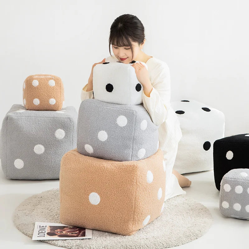 A person is holding a white polka-dotted plush dice cube cushion next to various gray and beige fabric cushions.