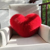 A bright red plush heart-shaped fox fur throw pillow resting on a light-colored armchair.