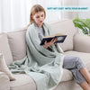 A woman is sitting on a sofa wrapped in a light green muslin throw blanket while reading a book.