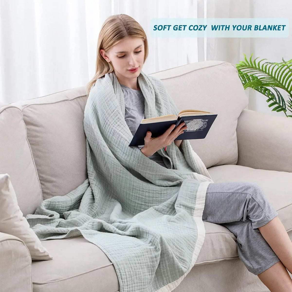 A woman is sitting on a sofa wrapped in a light green muslin throw blanket while reading a book.