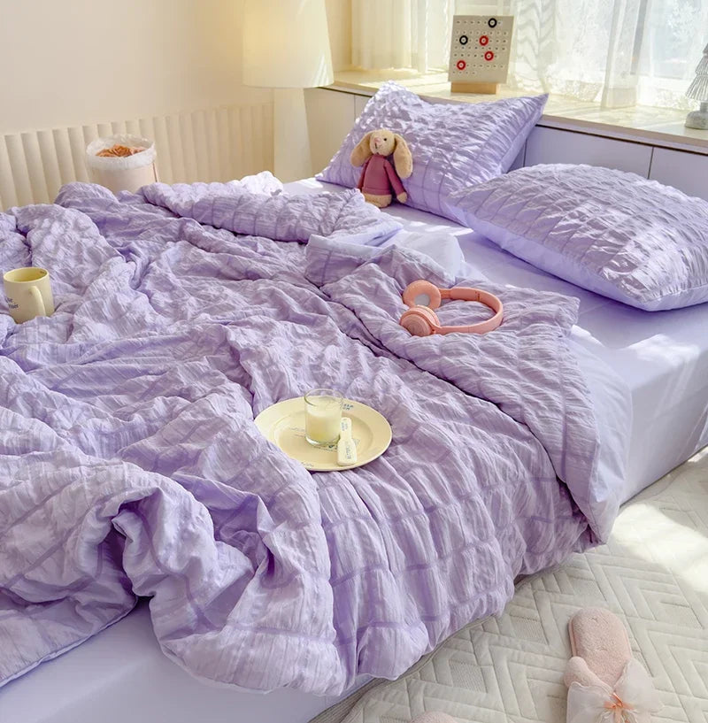 A purple summer seersucker quilt comforter spread on a bed with a white headboard and natural light.
