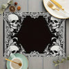 A black square placemat featuring a white gothic floral design with skulls and leaves, set on a wooden table.