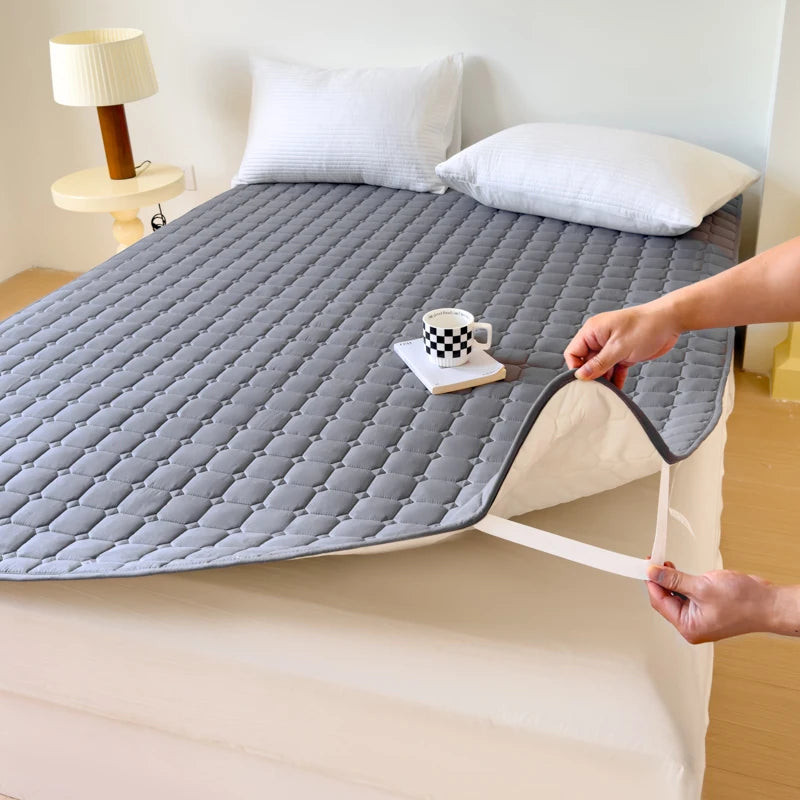 A gray quilted waterproof mattress protector is shown on a bed, with pillows and a mug placed on top.