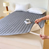 A gray quilted mattress protector cover is shown on a bed, with pillows and a mug placed on top.