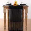 Black cheesecloth table runner draped over a wooden table with yellow flowers and glassware.
