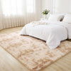 A fluffy cream tie-dye shaggy rug placed on a light wood floor next to a white bed and sheer curtains.
