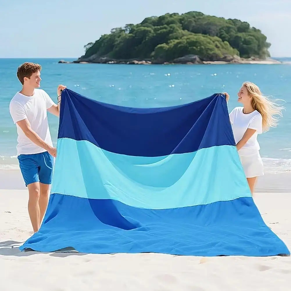 A large, folded blue and teal waterproof outdoor picnic mat is spread on a sandy beach with two people standing nearby.
