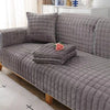 A gray quilted velvet sofa protection cover with decorative pillows and folded blankets on a wooden sofa.