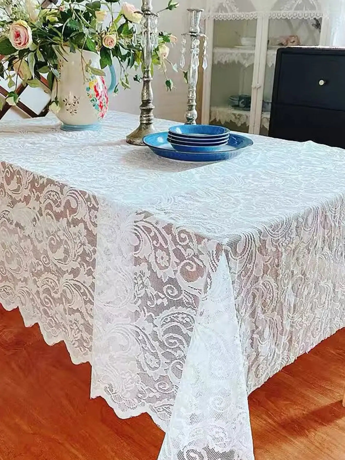 A white vintage lace tablecloth covers a rectangular dining table set with blue plates and floral arrangements.