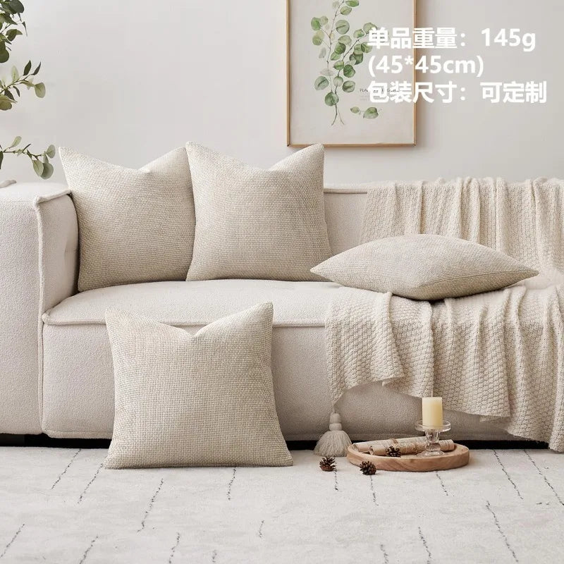 A beige chenille pillow cover displayed on a light-colored sofa with a sheer drape and a framed botanical print.