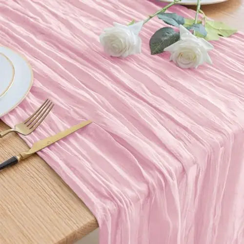 Pink pleated cheesecloth table runner with white roses and gold cutlery on a wooden table.