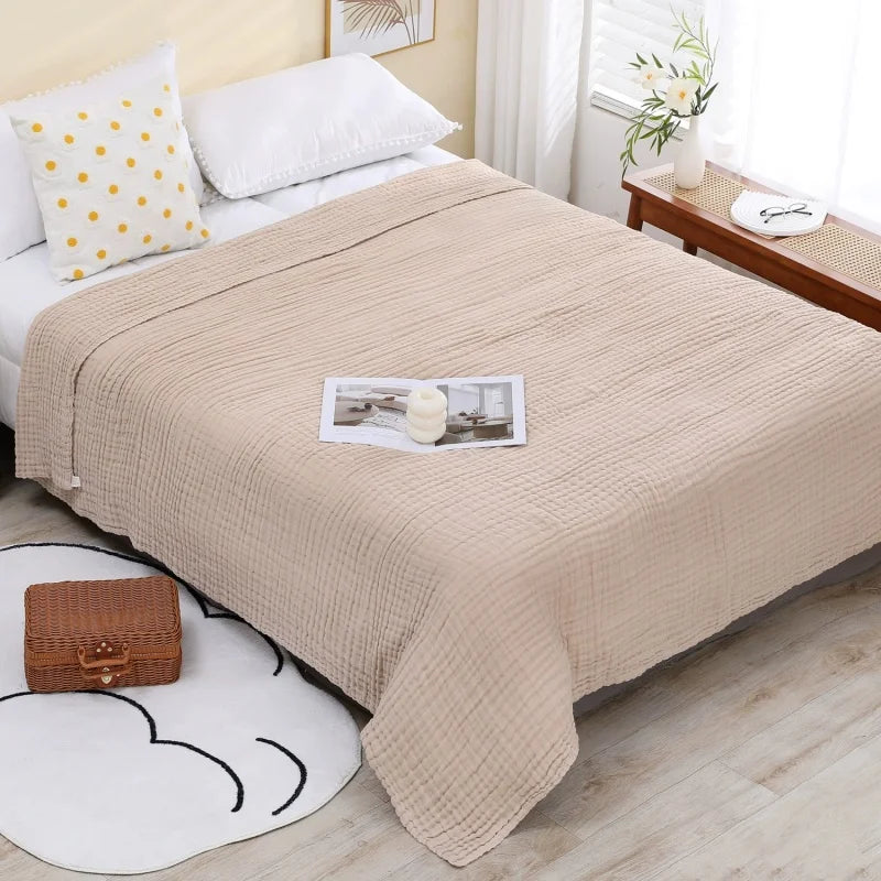 A beige pure cotton gauze throw blanket draped over a bed with white pillows and a wooden surface.