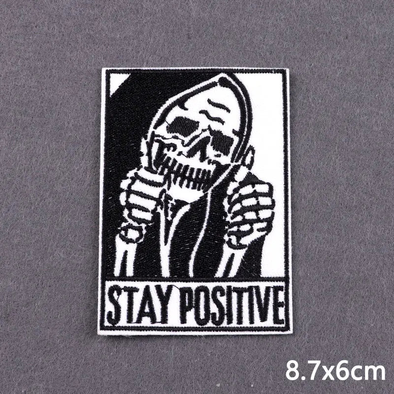 Black and white embroidered patch featuring a skull with hands to the face and the text STAY POSITIVE