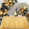 A gold sequin rectangle table cover is displayed on a table, surrounded by balloons and a cake.