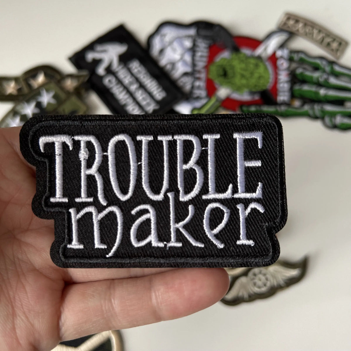 A hand holds a black iron-on patch with the white text TROUBLE maker, surrounded by other patches.