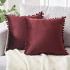 A deep maroon velvet decorative cushion cover with gold fringe resting on a white knitted throw.