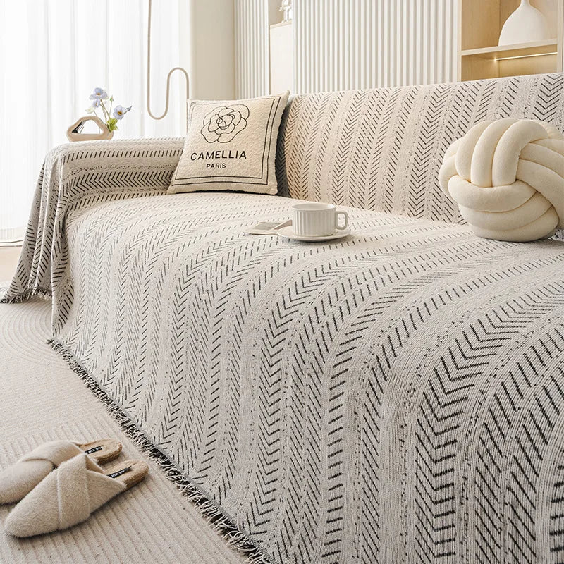 A beige and black fishbone pattern sofa cover with a cream pillow and a white cup on a light-colored bed.