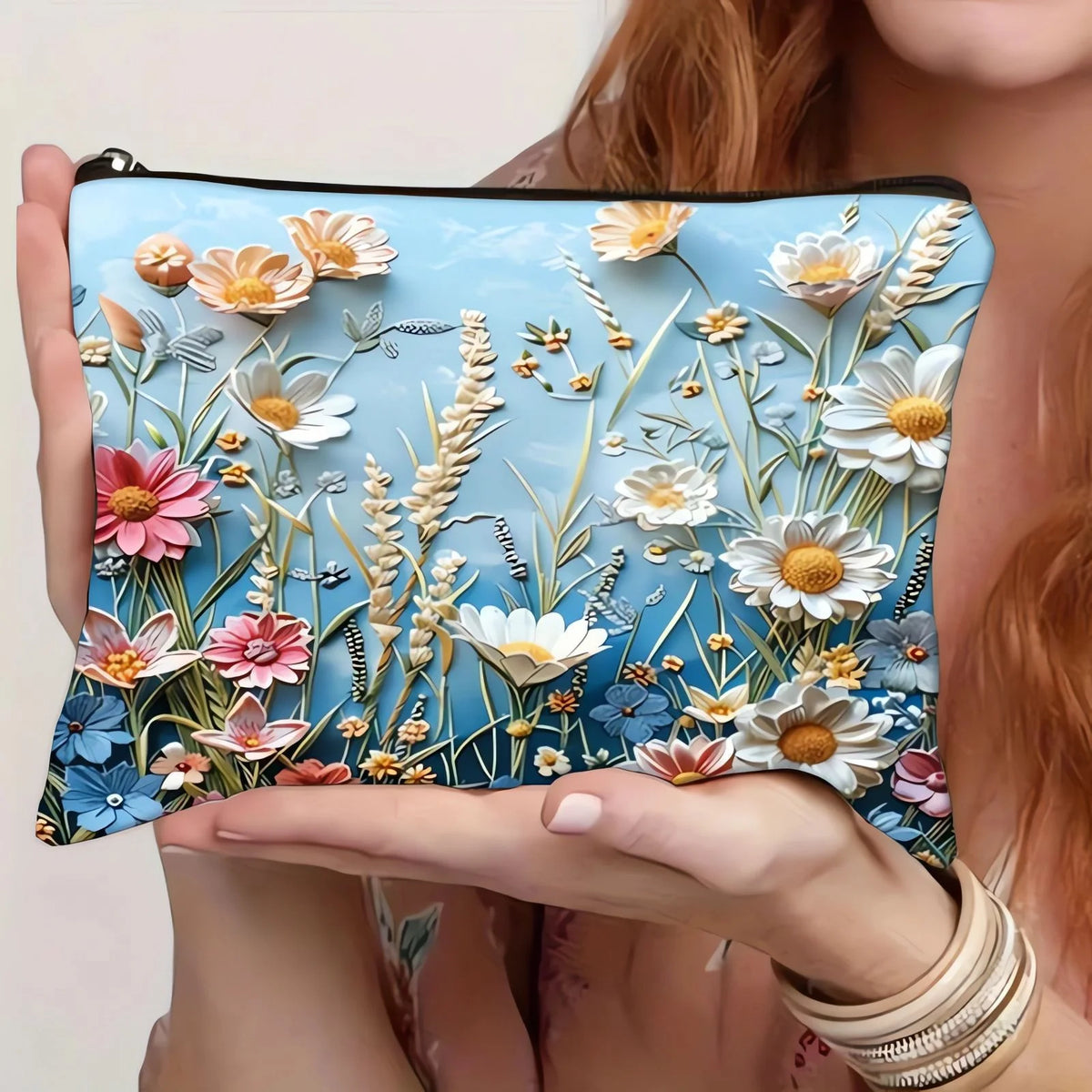 A hand holds a floral canvas cosmetic bag with a zipper closure featuring delicate white and pink flowers on a blue backgroun