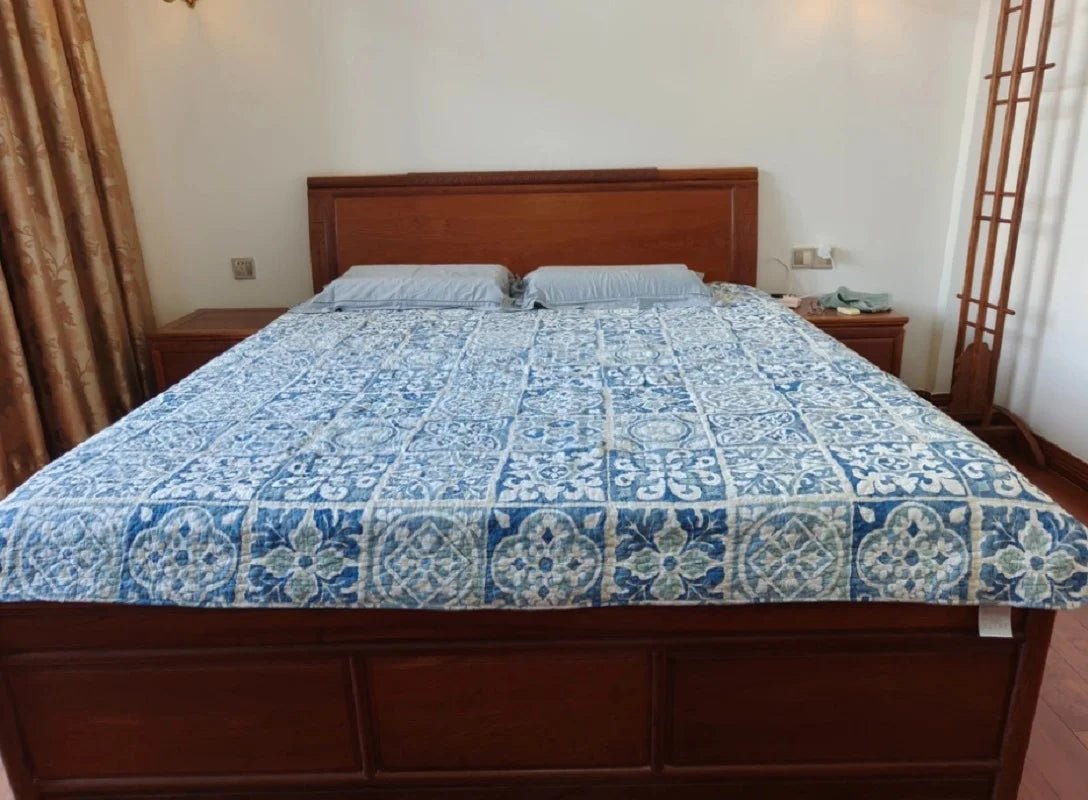 A wooden bed frame with a blue and white embroidered cotton quilted bedspread is shown in a bright bedroom.
