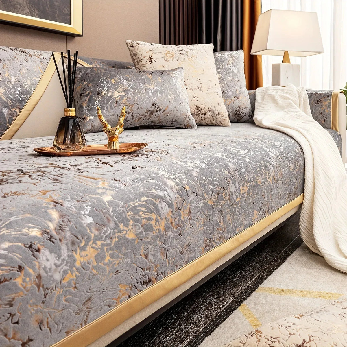 A luxurious gray velvet sofa cushion cover with gold foil detailing and a white throw blanket on a bed.