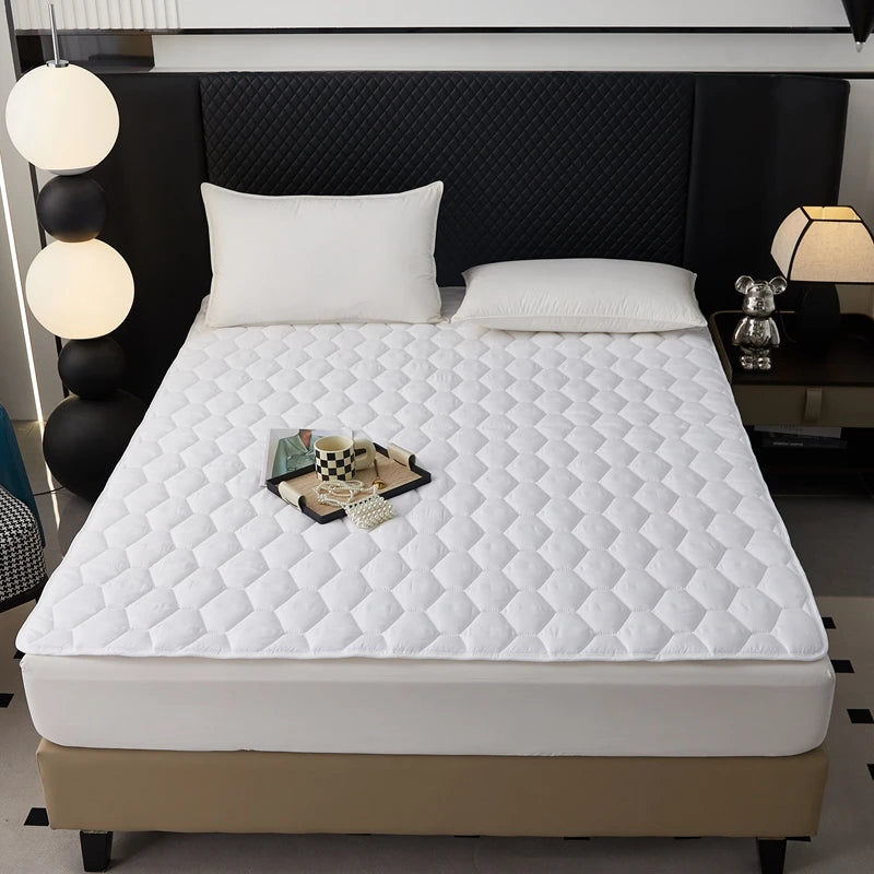 A white quilted mattress protector is displayed on a bed, featuring a diamond pattern and resting on a beige base.