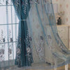 A dual-layer sheer blue curtain with embroidered floral patterns hangs in a bright room next to a light wood cabinet.