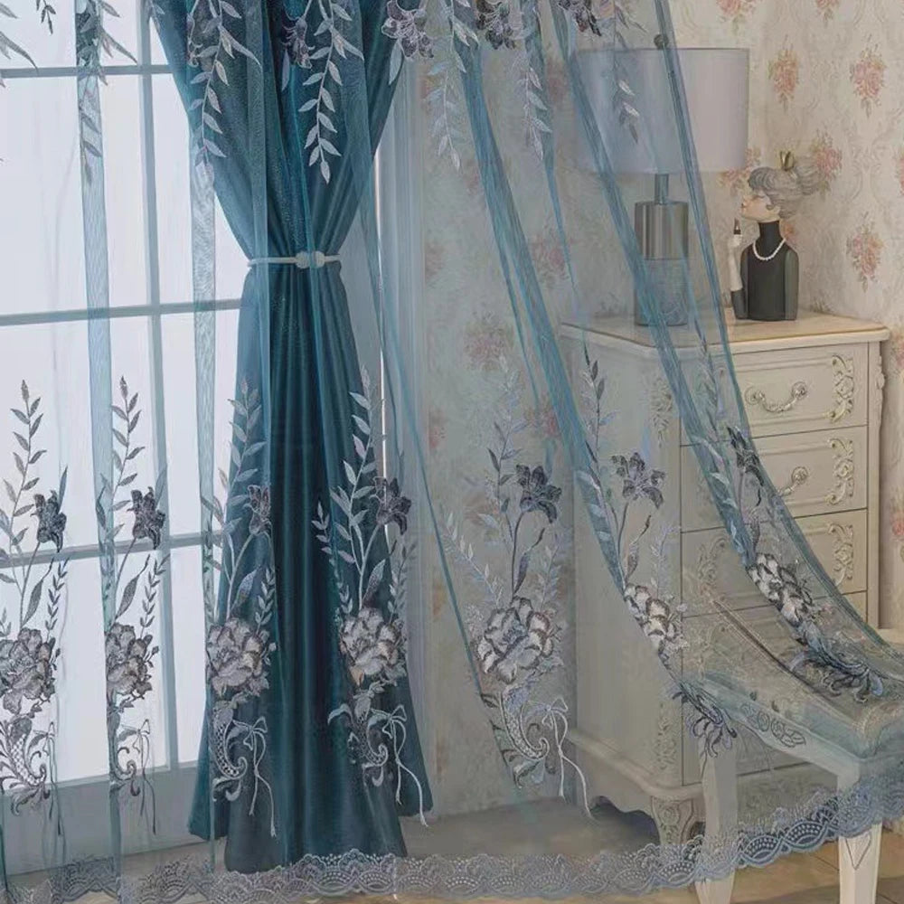 A dual-layer sheer blue curtain with embroidered floral patterns hangs in a bright room next to a light wood cabinet.