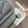 A hand is holding a white clip designed to tighten the corner of a bedsheet, shown next to a gray quilted fabric.