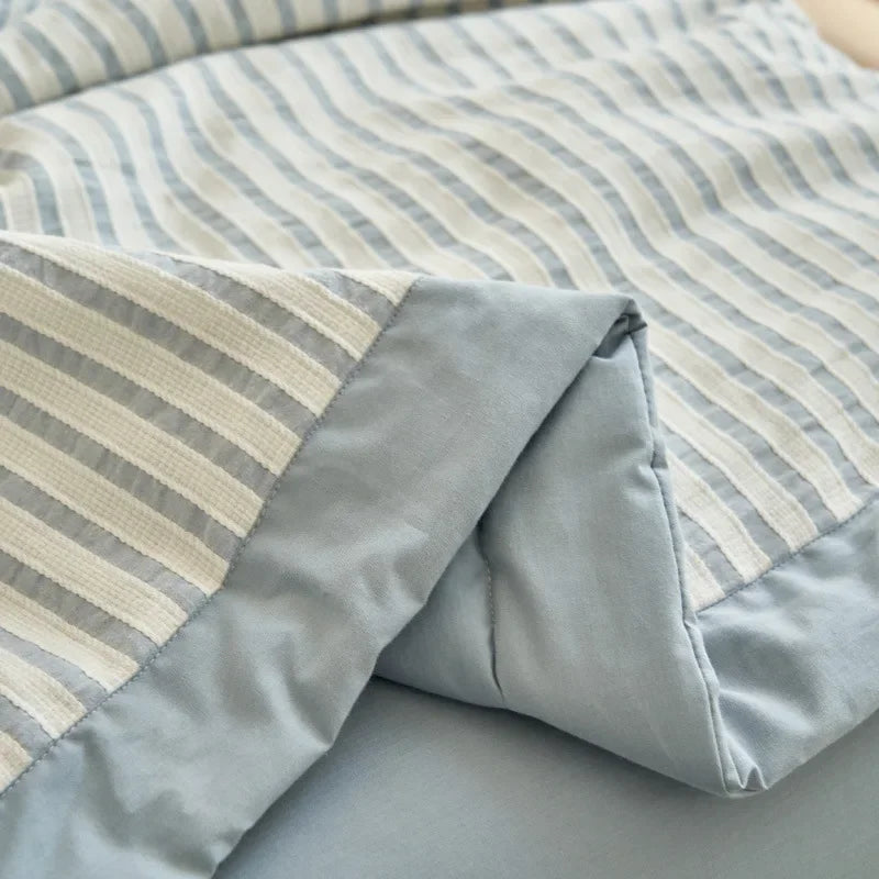A close-up of a light blue and white striped luxury jacquard summer quilt folded on a bed.