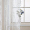 A sheer white floral lace curtain panel hanging in a window with a vase of white and blue flowers.