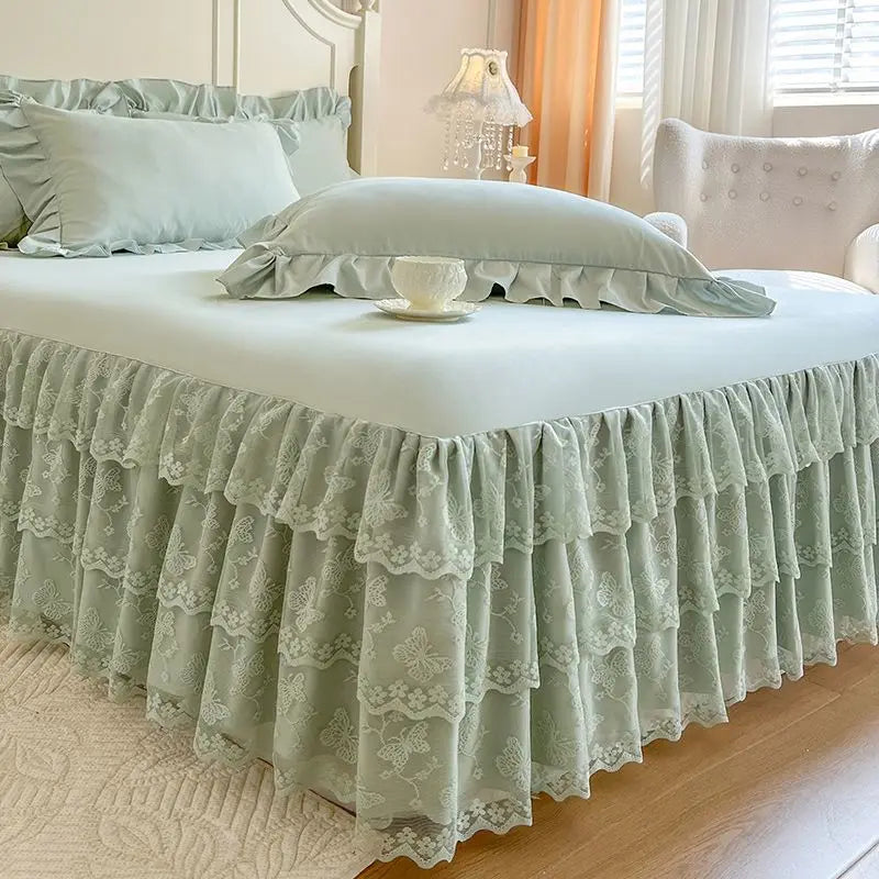 A bed dressed with a light green lace ruffled bed skirt and white linens in a bright bedroom.