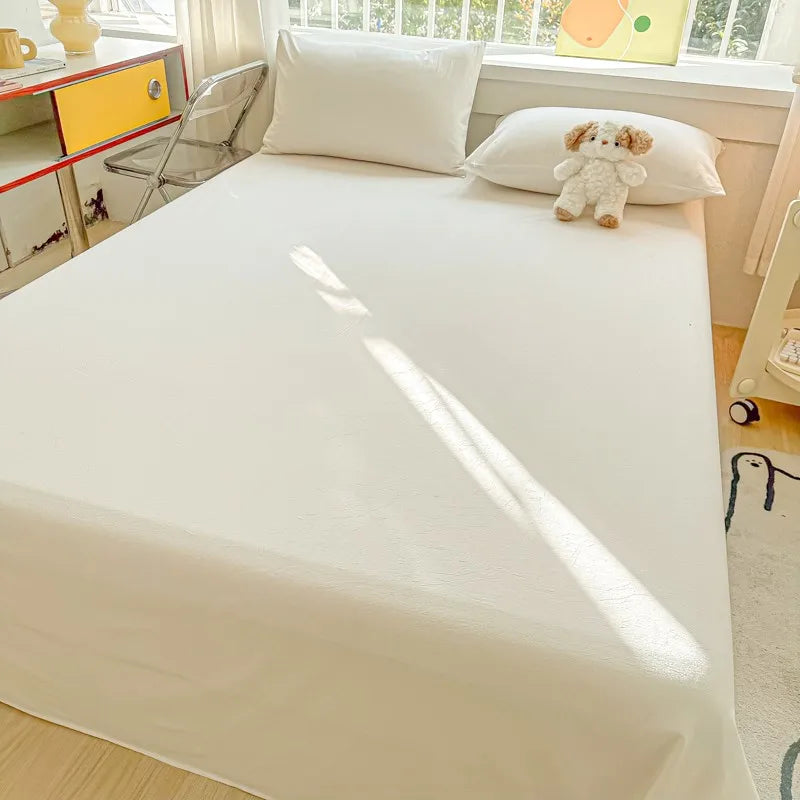A white organic cotton bedding set featuring a flat sheet, pillowcases, and a small teddy bear decoration on a bed.