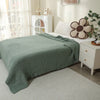 A light green quilted blanket draped over a white bed, next to a white nightstand with decorative items.