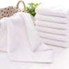 A stack of ten white cotton face towels is shown next to a folded towel on a white surface.