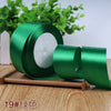 A roll of emerald green satin ribbon with white filling, displayed on a wooden surface with green foliage in the background.