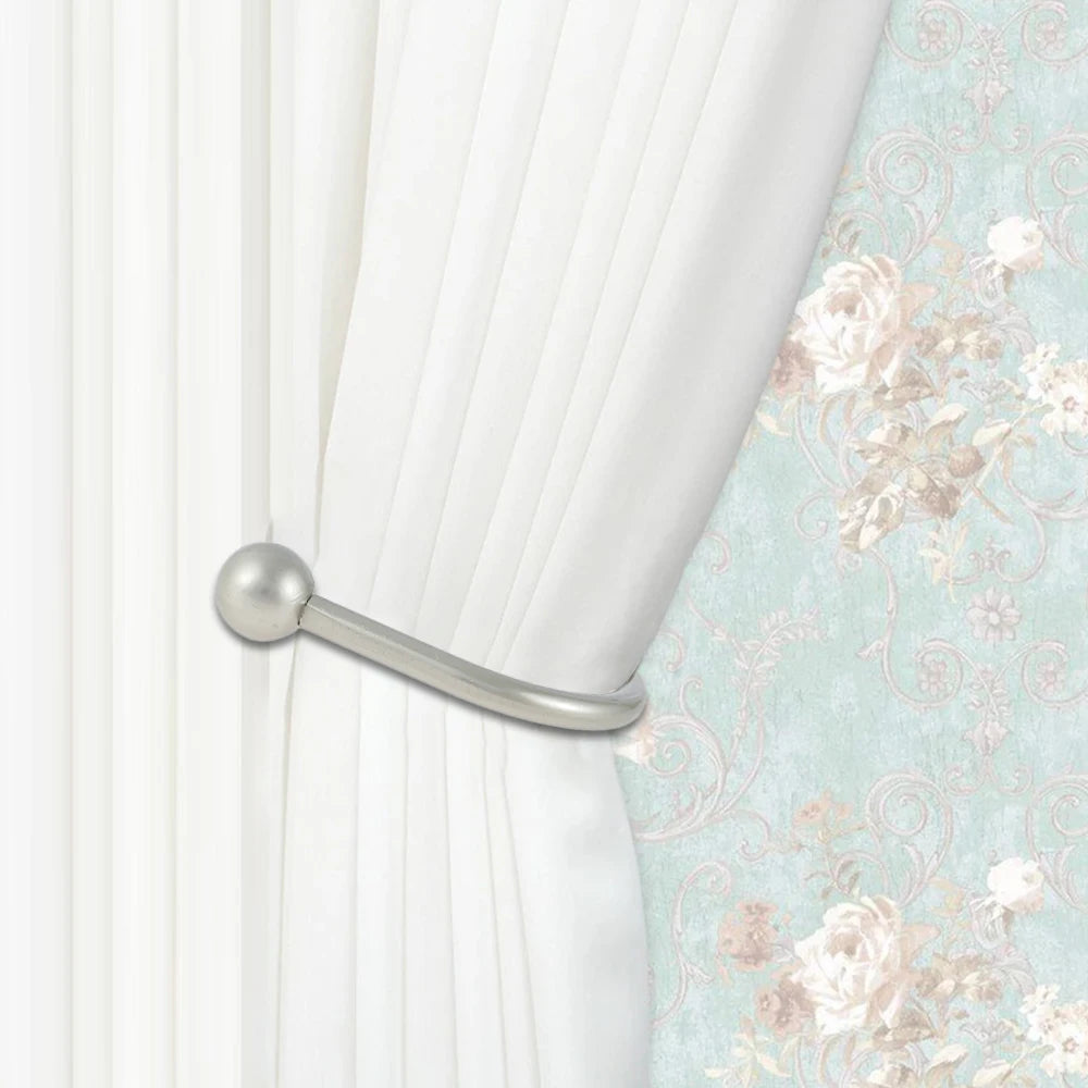 A silver metal curtain holdback hook is visible on a white pleated curtain against a light floral wallpaper.