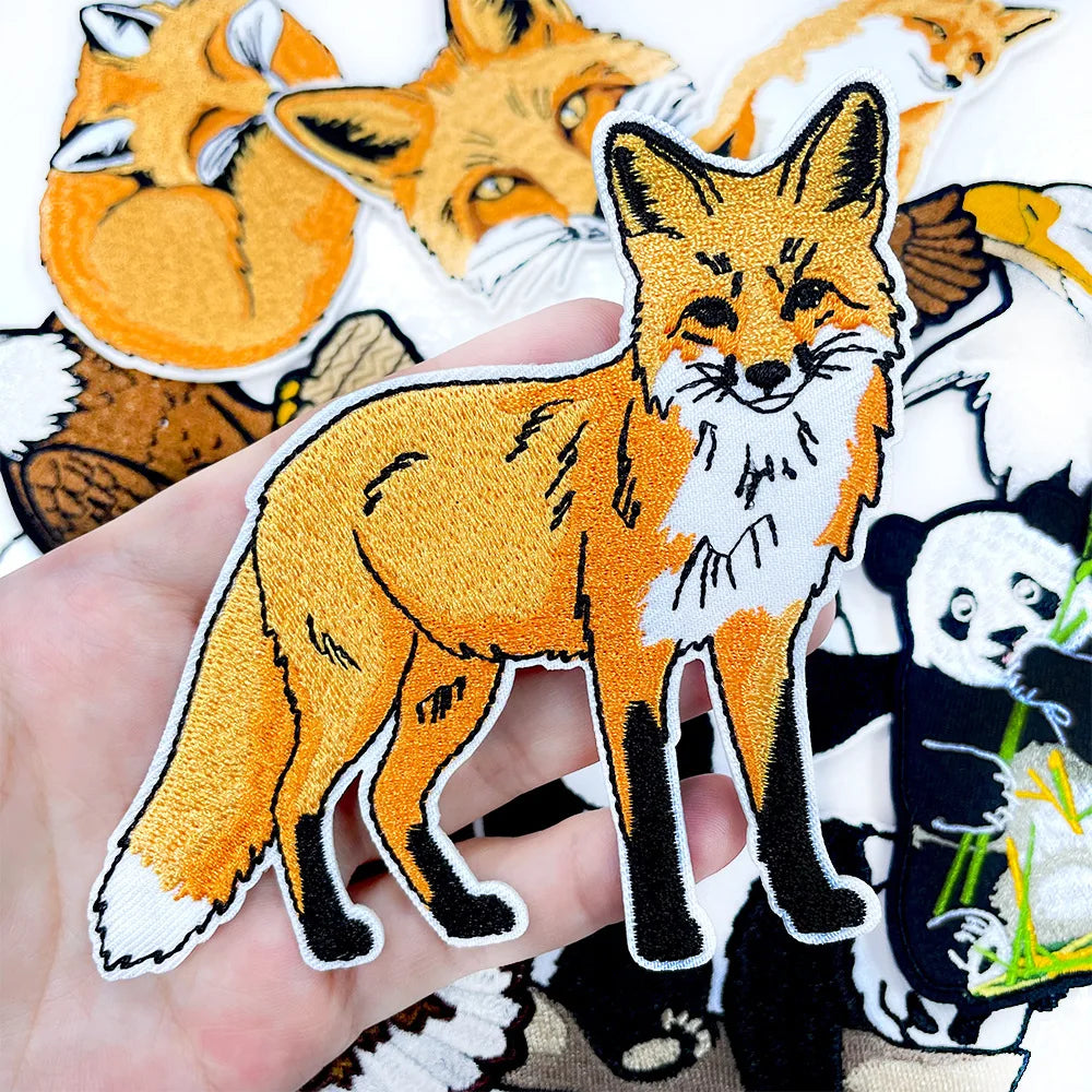 A hand holds an orange fox embroidery patch featuring white and black details.