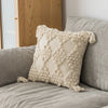 A beige plush pillow cover with a geometric pattern and fringed tassels resting on a gray sofa.