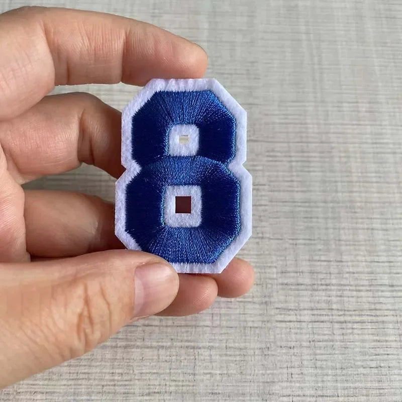 A hand holds a dark blue embroidered number patch with a white square inside on a light gray background.
