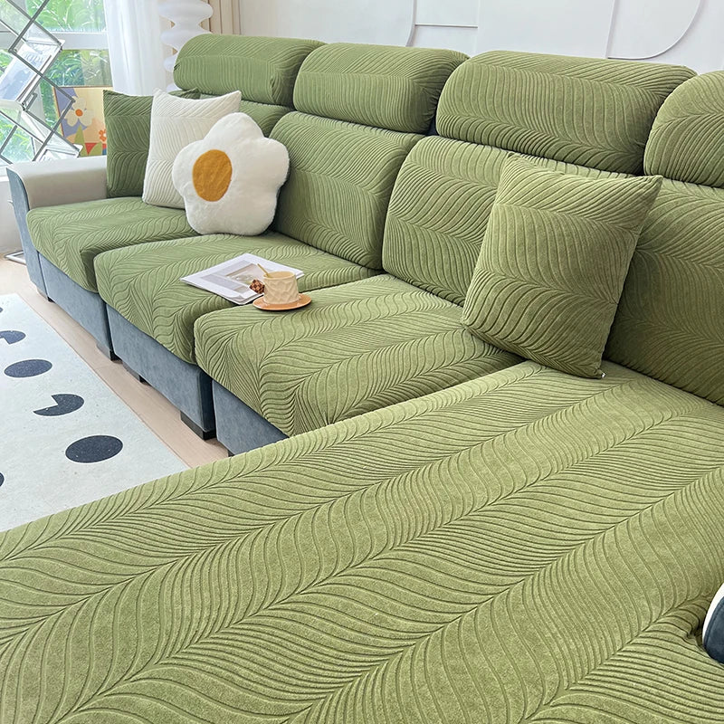 A sectional sofa covered in olive green jacquard stretch fabric with light cream accent pillows.