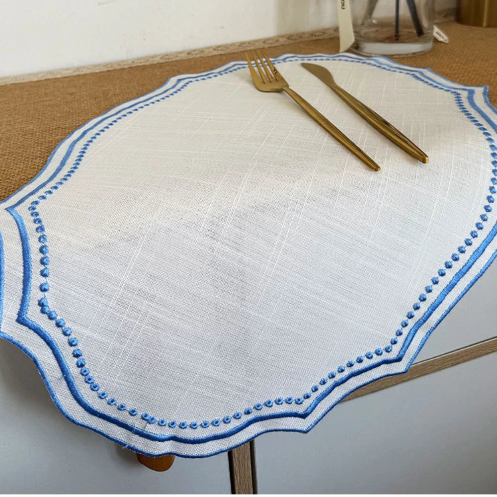 A white cotton linen placemat with blue embroidery and two gold-toned forks resting on it.