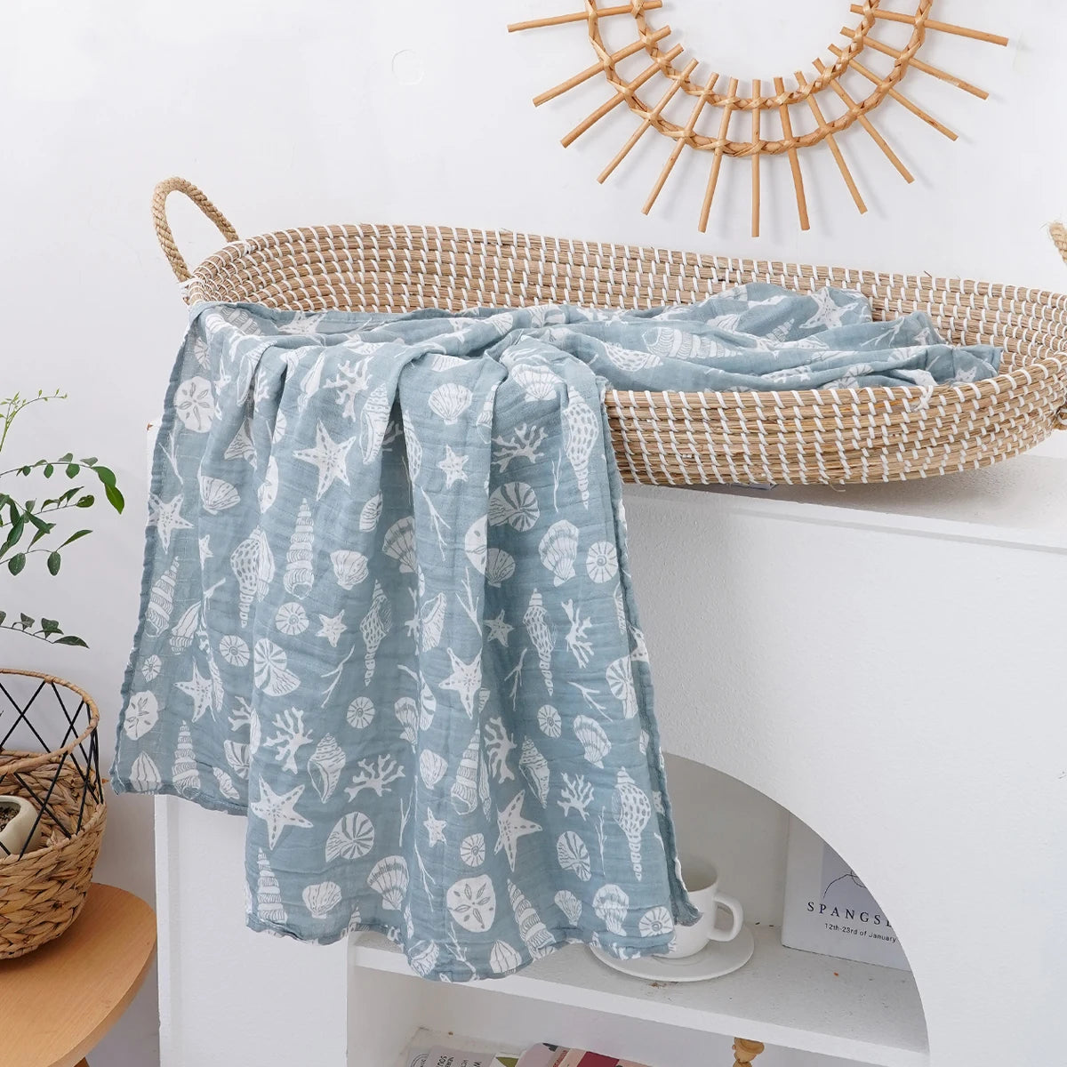 A light blue muslin swaddle blanket with a white leaf pattern draped over a woven basket on a white shelf.