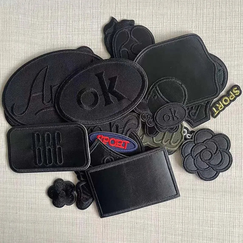 A set of five black embroidered leather patches with self-adhesive backing, including letters, symbols, and words like OK and