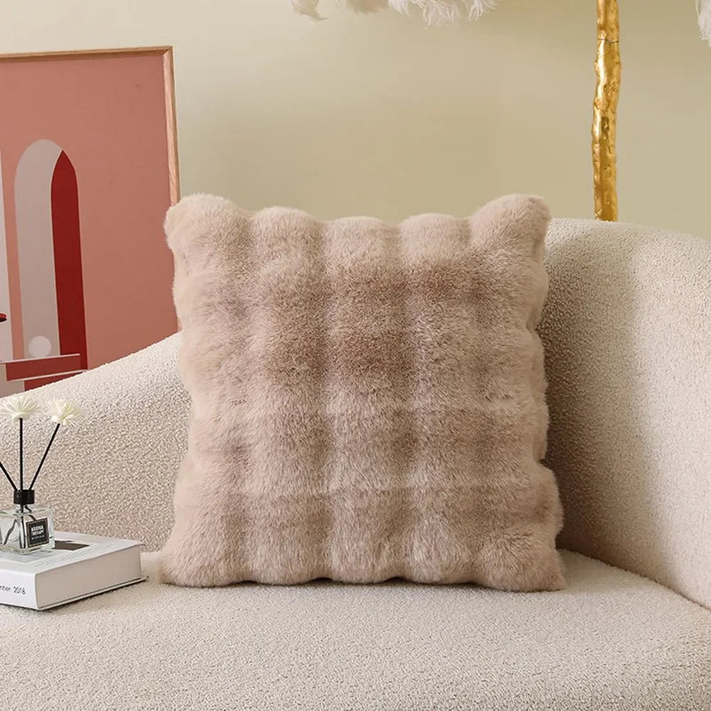 A square, light beige plush cushion cover with a textured, fluffy appearance resting on a cream-colored sofa.